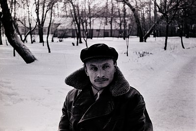 Portrait of a man in winter attire—fitted cap, fur-lined coat, and scarf—standing on a snow-covered path in a leafless park. ...