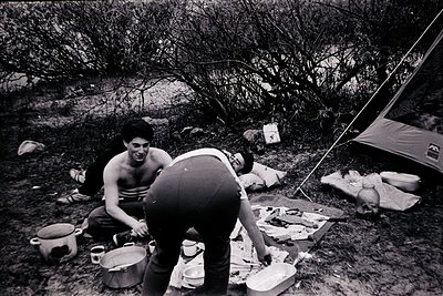 Mid-20th century camping scene with three individuals preparing food outdoors. Tent and camping gear visible in wooded, rocky...