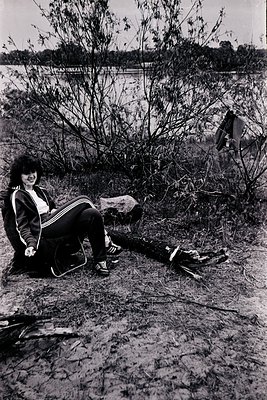 Black-and-white portrait of a woman in 1970s athletic wear—track pants, striped jacket, and sneakers—sitting on a fallen log ...