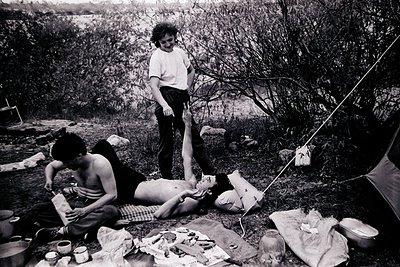 Black-and-white snapshot of a mid-20th-century camping scene. Three individuals—two adults and a child—relax by a stream in a...