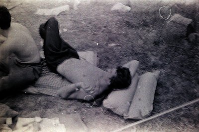Black-and-white photo of two individuals seated on concrete, likely mid-20th century. One person reclines on a folded blanket...