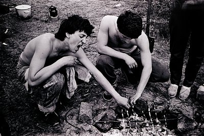 Black-and-white candid of two shirtless men building a small wooden structure over an open fire, likely a grill or cooking se...
