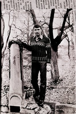 Young man in a patterned sweater poses beside a stone column in a wooded area, likely Eastern Europe, 1960s–1970s. Overlaid t...