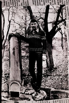 Young man in patterned sweater poses beside a brick chimney stack in a wooded, rural setting. Snow patches and bare trees sug...
