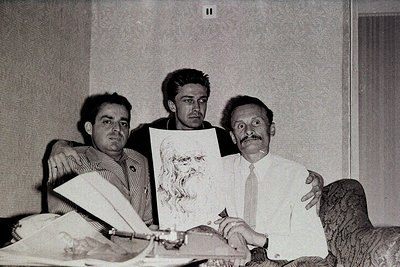 Three men pose indoors, mid-20th century (likely 1960s–70s). The seated man holds an abstract painting with bold, gestural br...