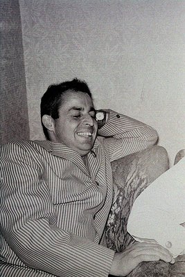 Portrait of a man in a striped button-down shirt, leaning back in a relaxed pose with one arm resting on a worn, textured sur...