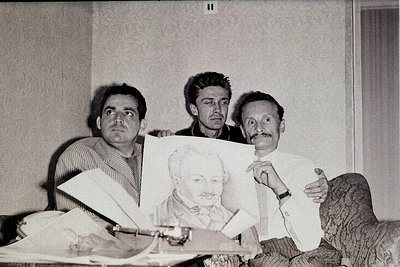 Black-and-white portrait of three men in a studio setting, mid-20th century. The man on the right holds a portrait of a beard...