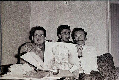 Three men pose indoors, holding a portrait of a bearded man in a suit. The setting appears to be a modest, dimly lit room wit...