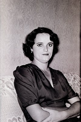 Portrait of a woman in a 1940s-style dress with a deep V-neck and rolled sleeves, seated indoors against a patterned wall. He...