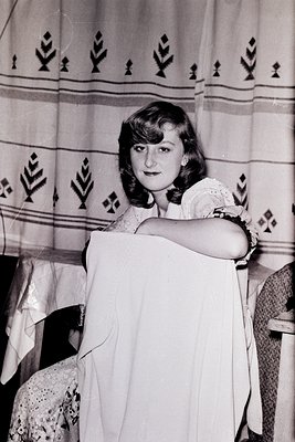 Young woman in 1950s-60s European folkwear, holding a white lace blouse against patterned embroidered fabric with geometric a...