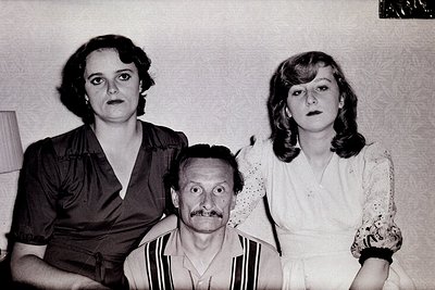 Black-and-white portrait of three individuals in formal attire, likely 1970s. Woman left wears a fitted blouse with a high co...