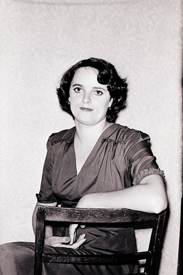 Portrait of a woman in a 1940s–1950s dress with a draped, off-shoulder neckline and short, voluminous hairstyle. Plain wooden...