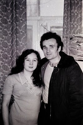 Black-and-white portrait of a man and woman posing indoors, likely mid-20th century. The man wears a leather jacket over a co...