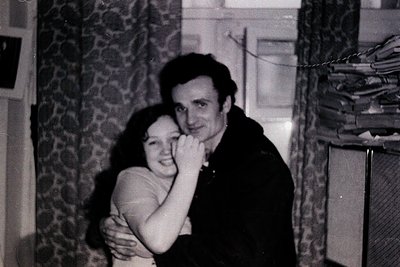 A tender indoor portrait from the 1970s–1980s, showing a man and woman embracing in a dimly lit room. The man wears a dark ja...