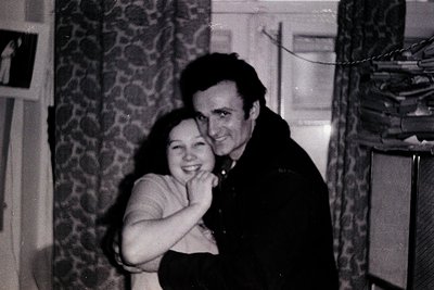 Black-and-white indoor portrait of a man and woman embracing, likely 1960s–1970s. The woman wears a sleeveless dress; the man...