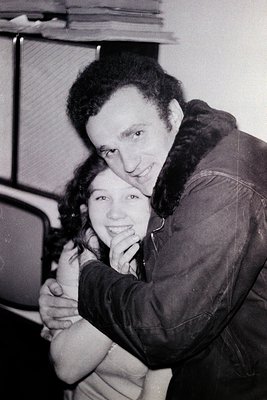 Black-and-white portrait of a man and woman embracing indoors, likely mid-20th century. The man wears a voluminous jacket and...