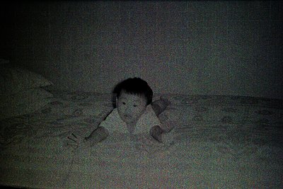 Vintage black-and-white photo of a child lying on a textured, patterned surface, possibly a bed or sofa. The low-angle shot e...