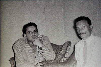 Black-and-white portrait of two men in formal attire, seated side-by-side. Left: man in striped shirt, chin resting on hand. ...