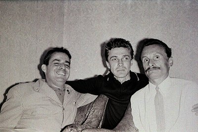 Three men pose casually in a mid-20th-century indoor setting, likely 1950s–1960s. Left: man in striped shirt, relaxed posture...