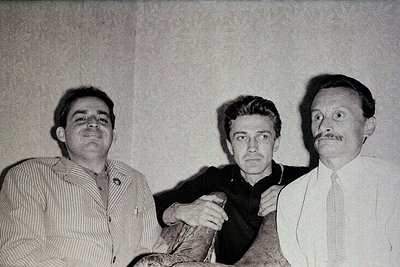 Black-and-white portrait of three men in mid-20th-century attire: striped shirt, dark blazer, and formal tie. The central fig...