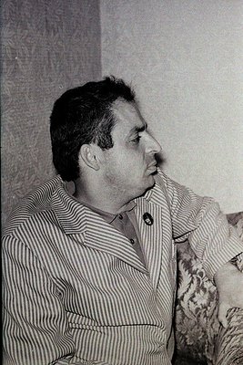 Mid-century man in striped button-up shirt, seated indoors against neutral wall. Classic 1950s–1960s hairstyle and attire sug...