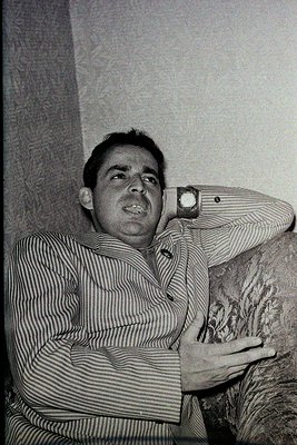 Black-and-white portrait of a man in a striped button-down shirt, leaning against a textured wall. His posture suggests casua...