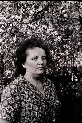 Portrait of a woman in a floral-patterned blouse, standing outdoors against a backdrop of blooming trees. Her hairstyle and a...