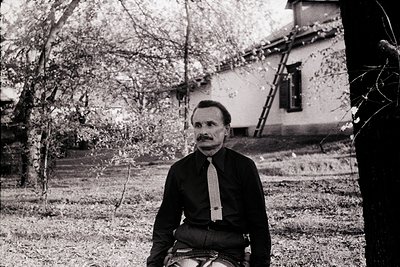 Black-and-white portrait of a man seated on a motorcycle in a rural outdoor setting, likely mid-20th century. He wears a dark...