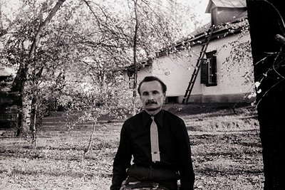 Mid-century man in formal attire poses outdoors beside weathered wooden house. Dark tie and suit suggest 1950s–1960s European...