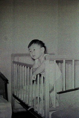 Vintage black-and-white photo of a young child leaning on a wooden crib railing, dressed in a short-sleeved dress with a bow....