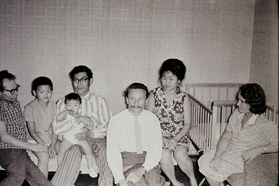 Family portrait from the 1970s, likely indoors in a residential setting. Five adults and a baby pose in a living room with a ...