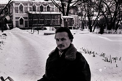 Mid-century man in winter coat poses on snow-covered path beside historic Gothic-style building with pointed arches and stain...