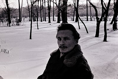 Portrait of a man in a winter park setting, likely mid-20th century. He wears a fur-lined coat and a mustache, standing on sn...