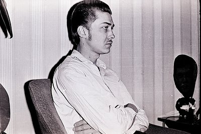 Portrait of a young man in a 1960s-70s indoor setting, seated with arms crossed. Light-colored button-down shirt, short hair,...