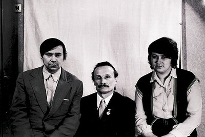 Three individuals pose formally in a 1970s indoor setting, likely Eastern Bloc. Center figure wears a suit, tie, and medal—su...
