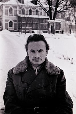 Portrait of a man in a winter coat with fur collar, standing on snow-covered ground. Gothic-style church with pointed arches ...