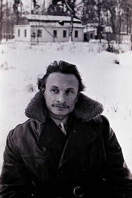 Mid-century man in a fur-collared coat poses against a snowy, rural backdrop. Snow-covered ground and a distant, weathered ho...