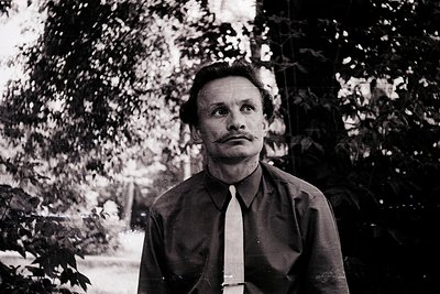 Portrait of a man in mid-20th century formalwear—dark suit, tie, and mustache—set against blurred greenery. Classic black-and...