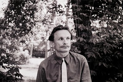 Portrait of a man in mid-20th-century attire—dark button-up shirt, tie, and neatly combed hair—standing outdoors against blur...