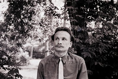 Portrait of a man in mid-20th century attire—dark suit, tie, and short hair—standing outdoors among blurred foliage. Classic ...