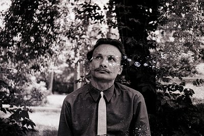 Portrait of a man in mid-20th century formalwear—dark suit, white tie, and mustache—standing outdoors under dense foliage. Bl...