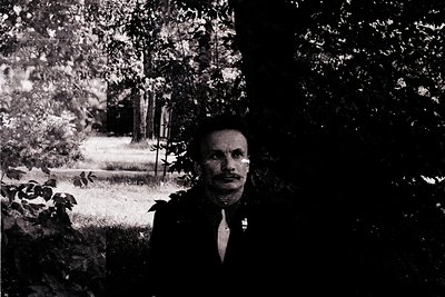 Mid-20th century black-and-white portrait of a man in formal attire—dark suit, white shirt, and tie—standing outdoors under d...