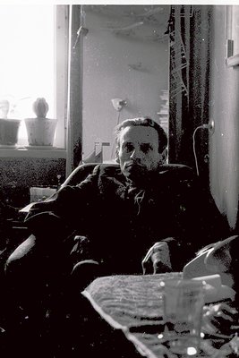 Mid-century man in a dimly lit room, seated on a sofa with a newspaper spread across his lap. The setting suggests a 1950s–19...