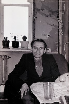 Mid-century man in a dark suit poses indoors beside a radiator, framed by a window with potted plants. The setting suggests a...