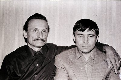 Two men pose closely in a candid black-and-white photo, likely from the 1970s–1980s. The elder wears a mustache and dark butt...