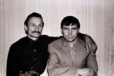 Two men pose closely in a candid black-and-white portrait, likely from the 1960s–1970s. The older man sports a mustache and a...