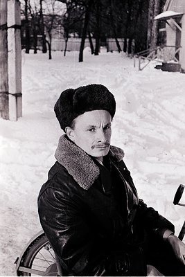 Mid-20th century man in winter attire—fur-lined coat, fur hat, and scarf—sitting on a snow-covered bench in a wooded park. Sn...