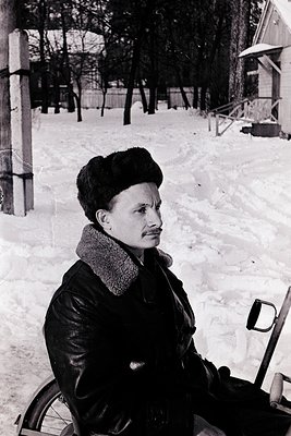 Mid-century winter portrait of a man in a leather jacket and fur-trimmed collar, seated on a snow-covered path. His fur hat a...