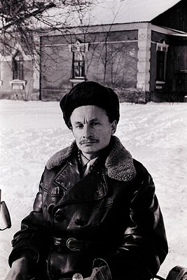Mid-20th century man in winter attire—leather jacket, fur collar, and flat cap—sitting on a snow-covered surface. Rustic bric...