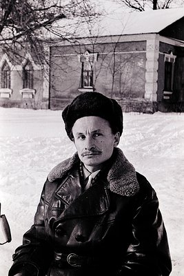 Mid-century portrait of a man in winter attire—leather jacket, fur collar, and beret—standing in snow-covered courtyard. Monu...